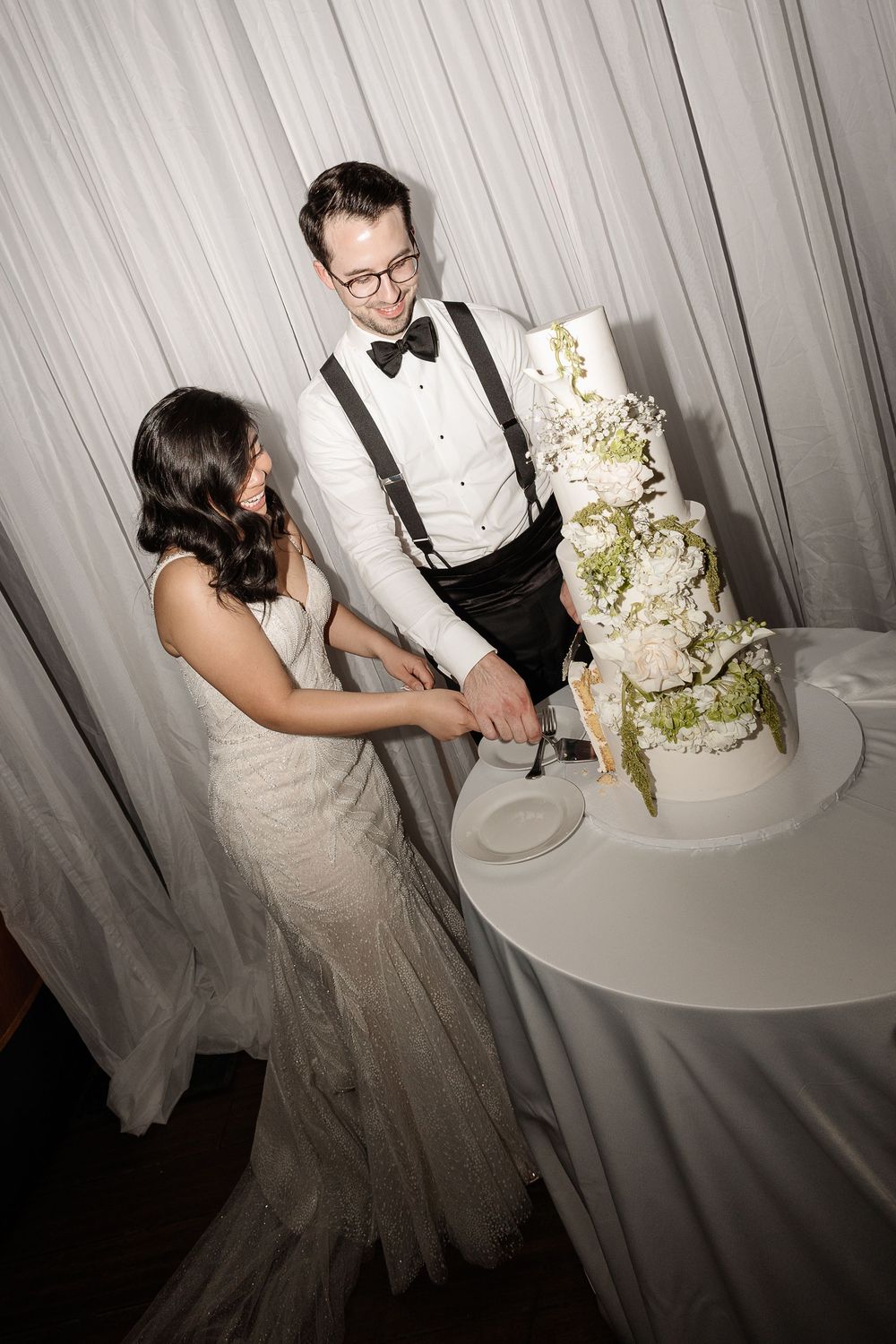 Smiling bride and groom cut tiered white floral wedding cake together at elegant reception.