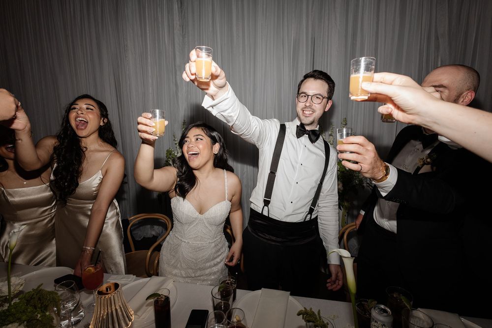 Bride and groom raise glasses in a joyful toast with wedding party at the reception table.