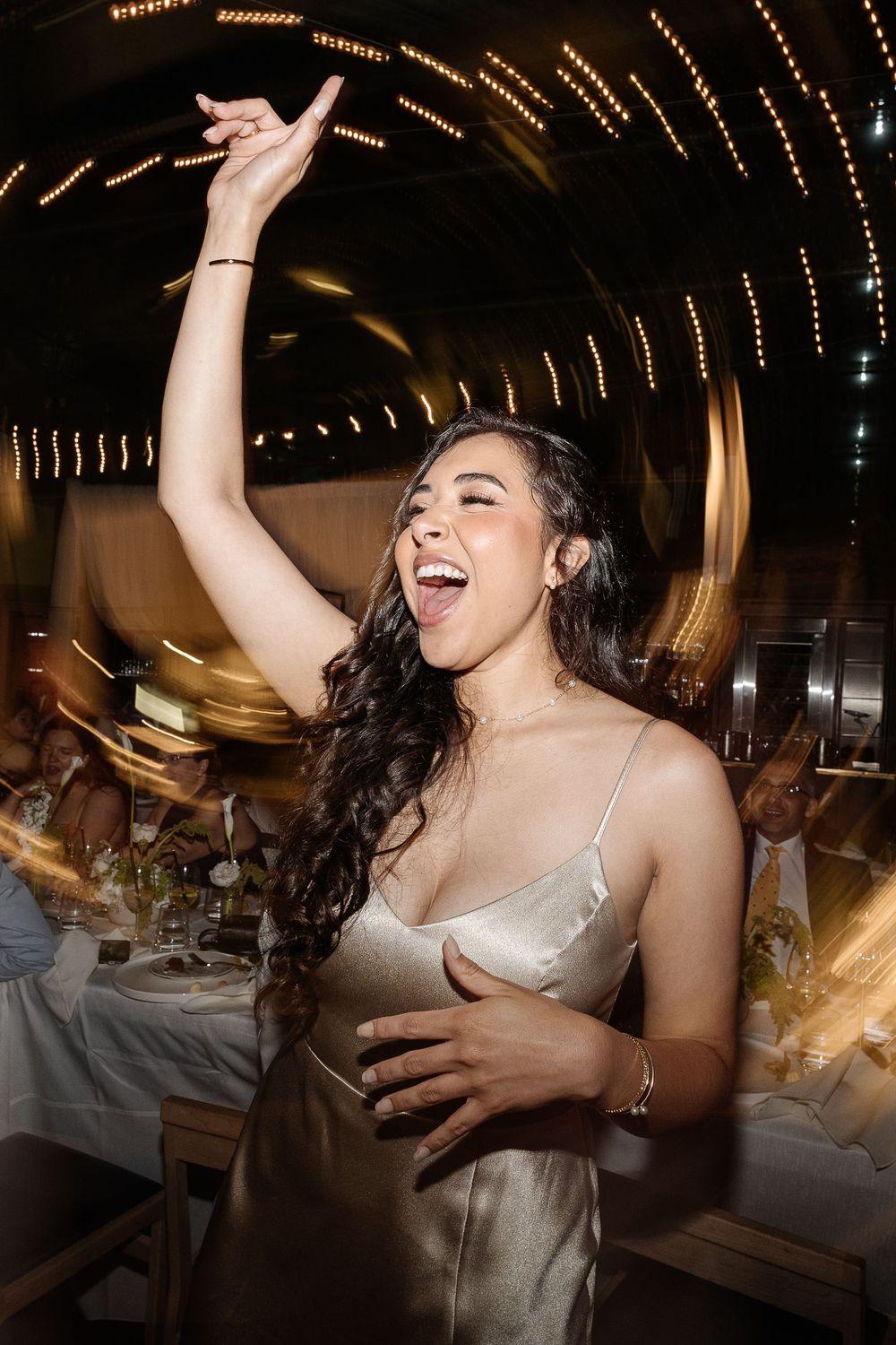 A joyful woman in a satin slip dress dances with arm raised at a warmly lit wedding reception.