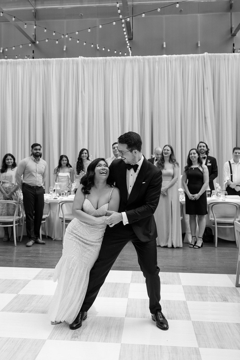 Bride and groom share their first dance on a checkered floor while wedding guests watch in the background.