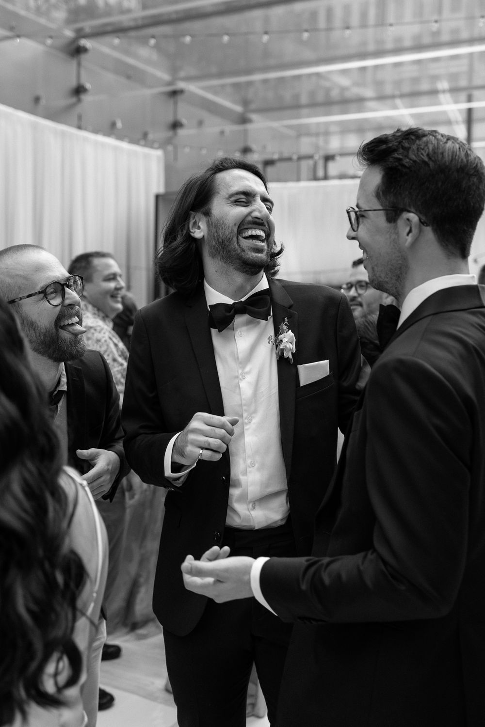 Laughing groom in black tuxedo and bow tie socializing with guests,  captured by Aliza Dworkind at Le Cathcart, a modern wedding venue.