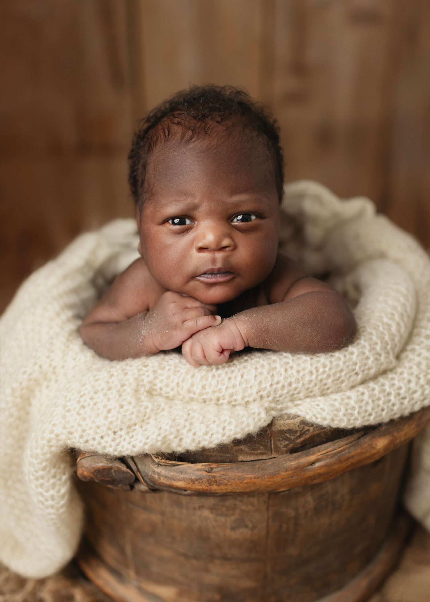 Newborn baby resting in a rustic wooden bucket wrapped in a cream knit blanket, gazing at the camera. Elizabeth Lang Photography Surrey