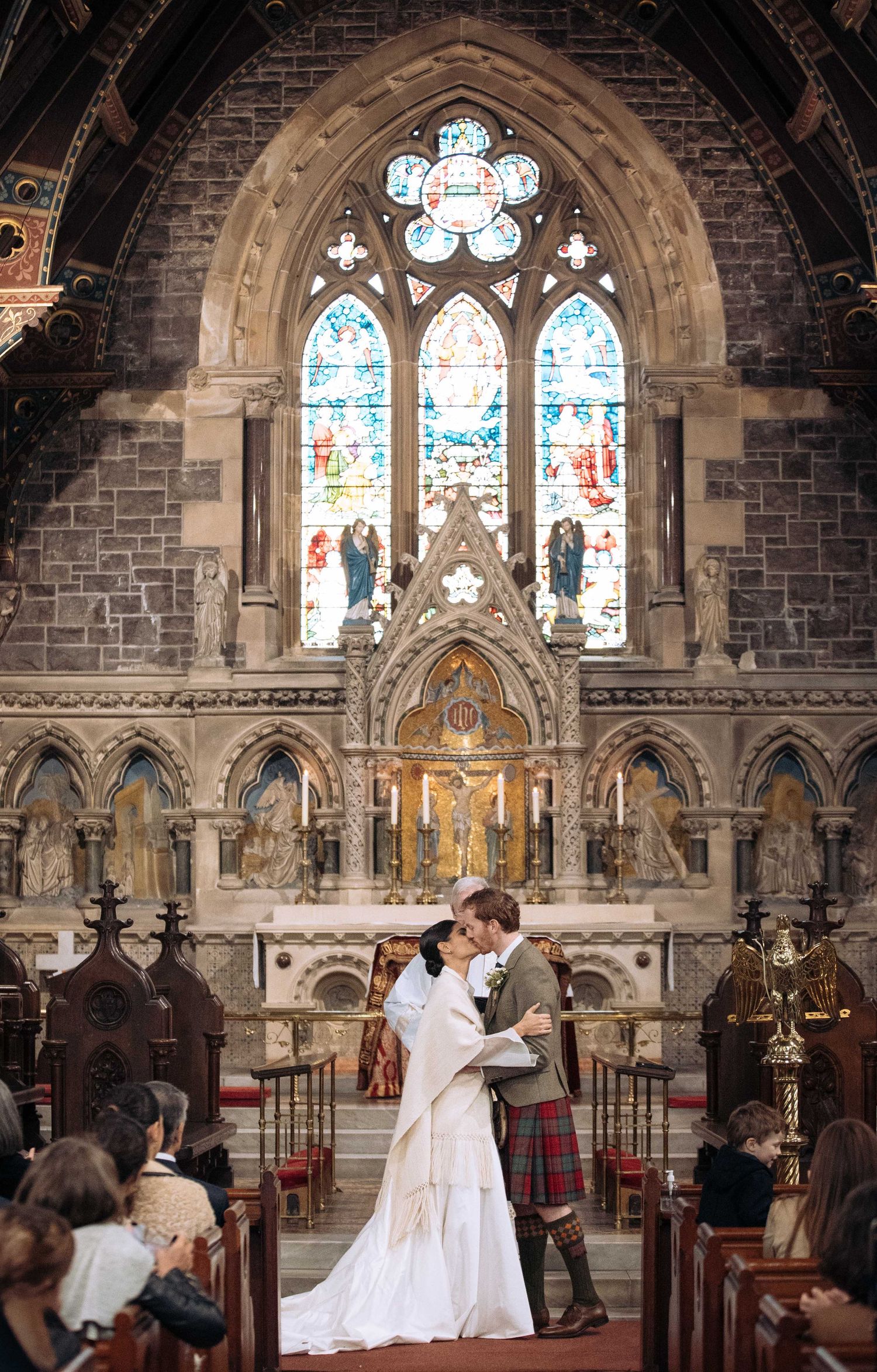 Bride and groom share first kiss at ornate church altar with stunning stained glass windows during Scottish wedding ceremony.