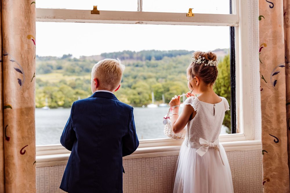 Two children in formal attire, a boy in navy suit and girl in white dress, gazing out a window at a scenic lake view.
