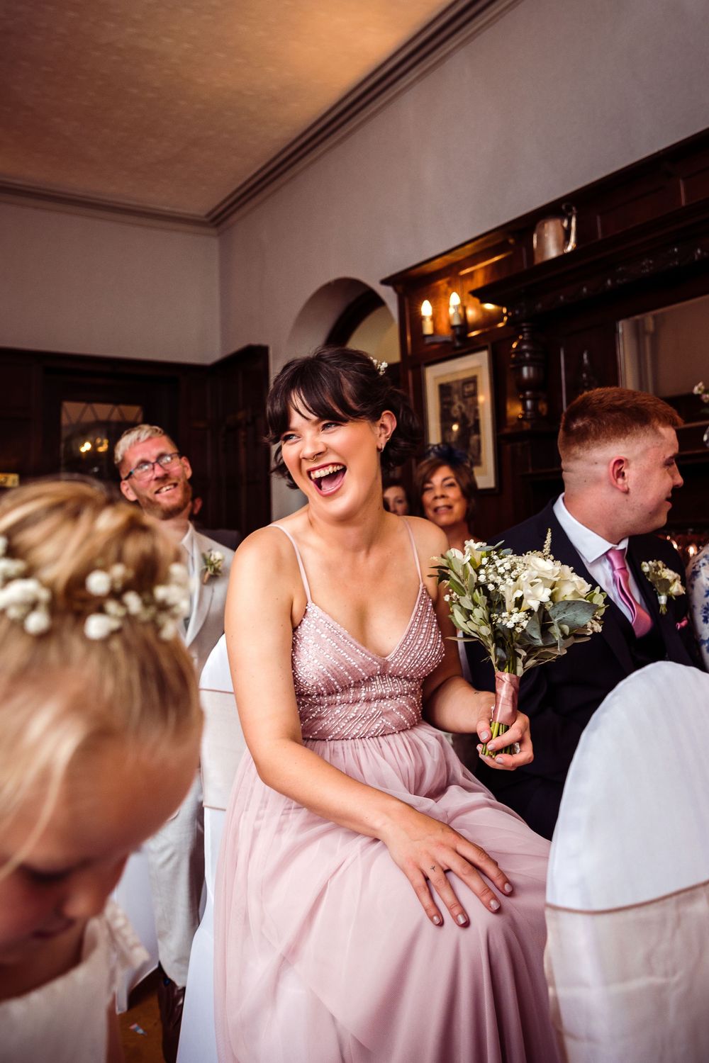 Laughing bridesmaid in pink dress holding white bouquet during wedding ceremony in rustic venue.