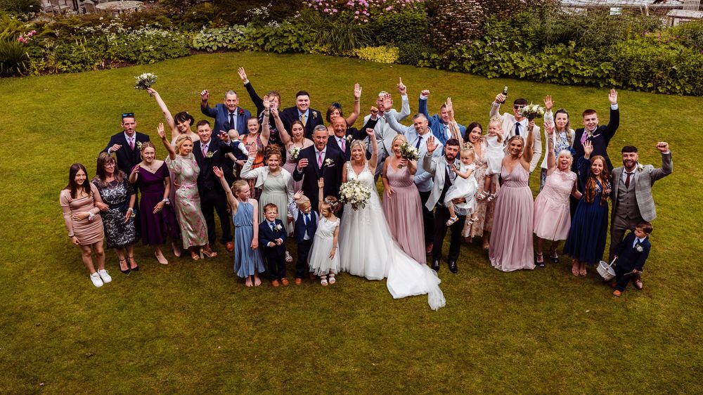 Large wedding party celebrating outdoors on green lawn, bride in white gown surrounded by guests in formal attire cheering joyfully.