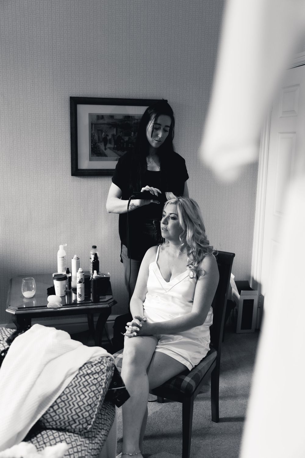 Bride getting hair styled by hairdresser in hotel room before wedding, black and white photo.