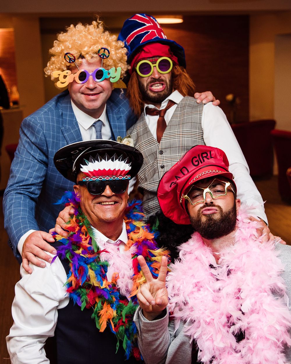 Four wedding guests posing in colorful costumes, wigs, and feather boas at a fun photo booth.