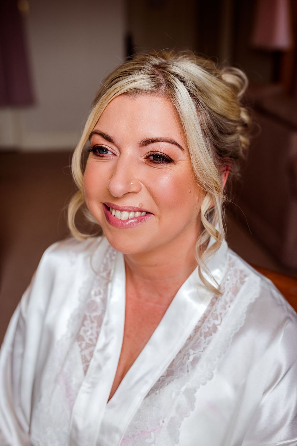 Smiling blonde bride in white satin robe with lace detail, wearing elegant updo hairstyle and natural bridal makeup.