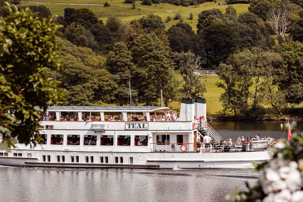 A white passenger cruise boat sailing on a calm lake surrounded by lush green hills and trees on a sunny day.