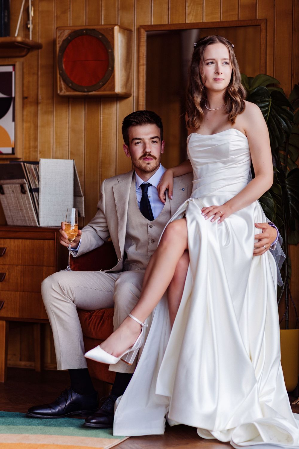 Elegant bride in white strapless gown and groom in beige suit posing together in a vintage-styled room with champagne.
