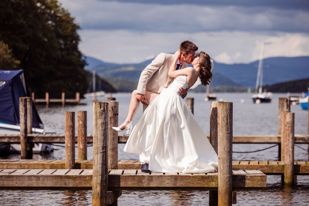 Bride and groom share a romantic dip kiss on a wooden dock overlooking a serene lake with sailboats.