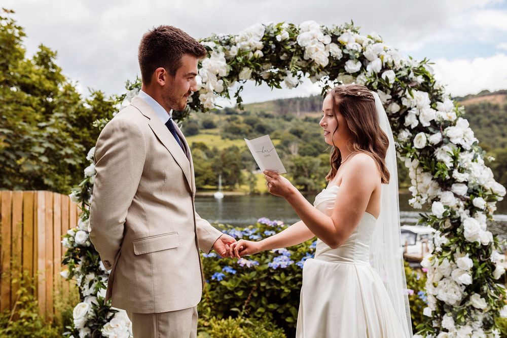 Bride reads wedding vows to groom under white floral arch outdoors by a lake.