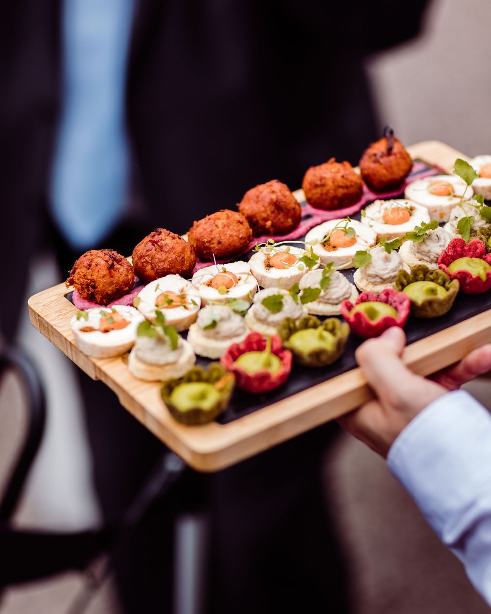 A server holds a wooden board with elegant canapes including fried balls, cream cheese bites, and colorful tartlets.