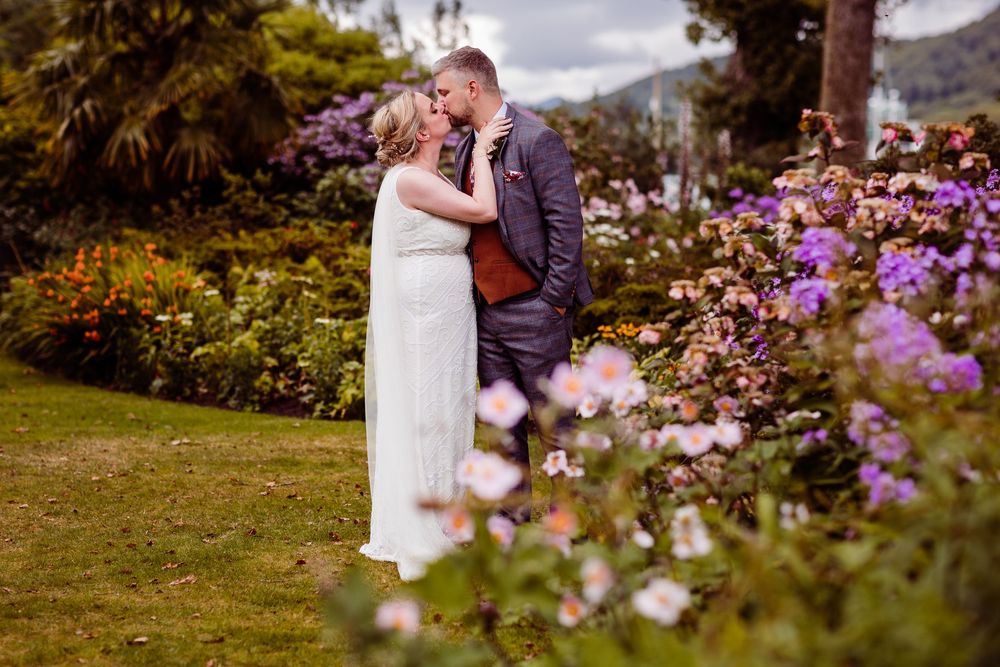 Bride and groom kissing in a lush garden surrounded by colorful purple and pink flowers on their wedding day.