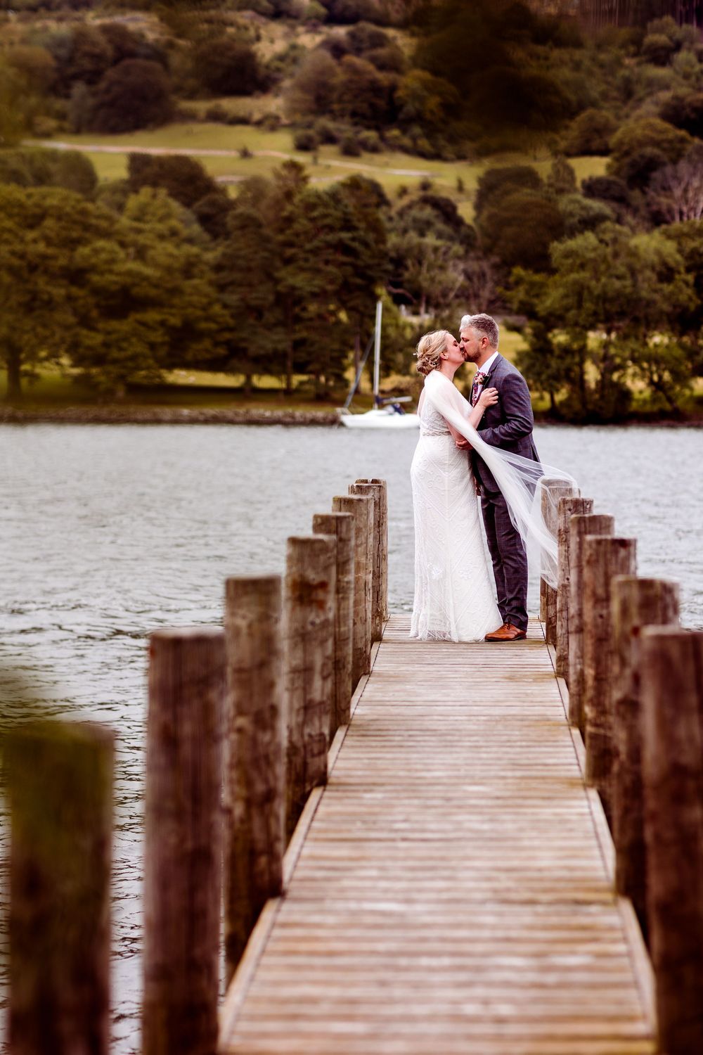 Bride and groom share a romantic kiss on a wooden pier over a calm lake with green hills in the background.