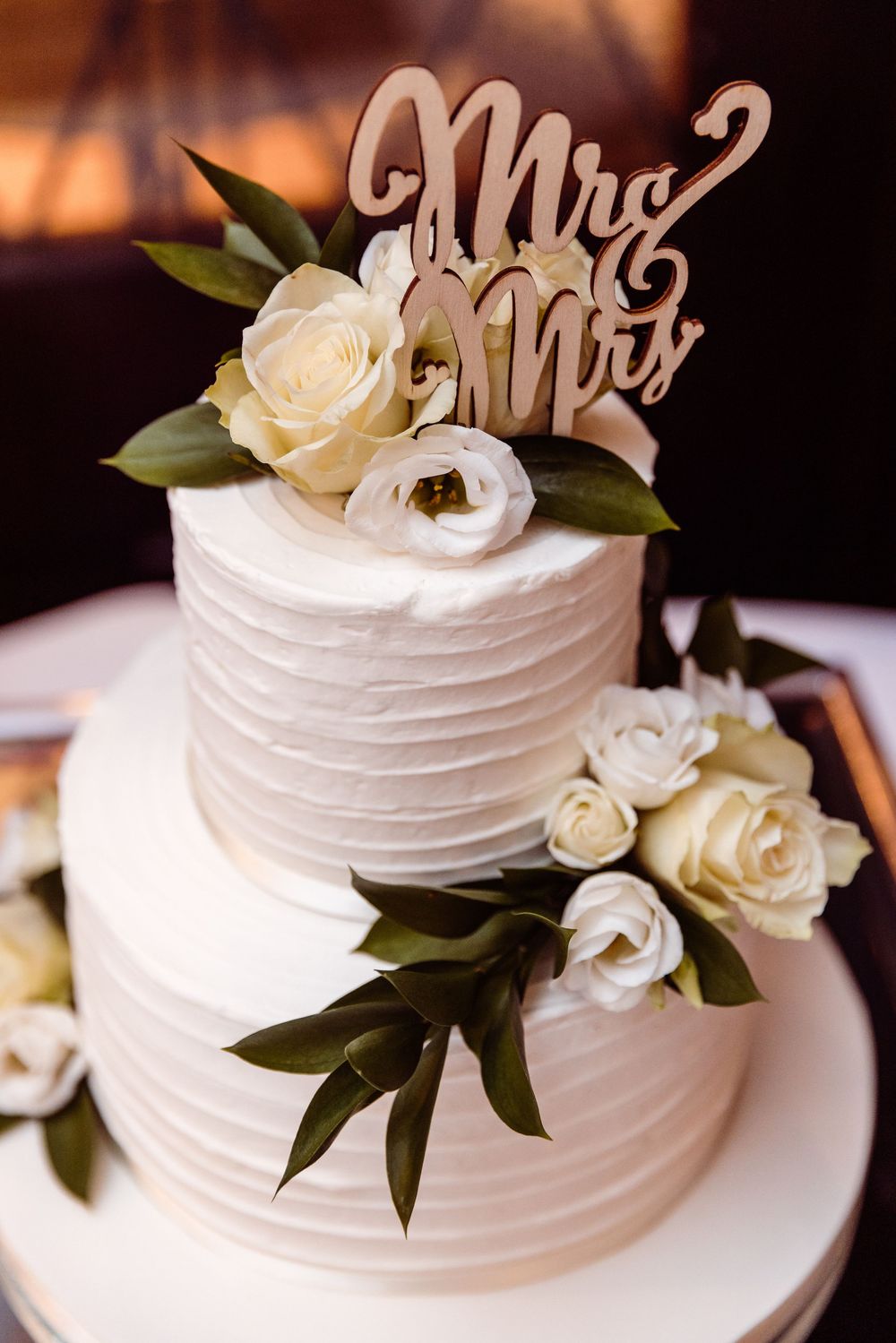 Two-tier white textured wedding cake with cream roses, greenery, and a wooden Mrs. & Mrs. cake topper.