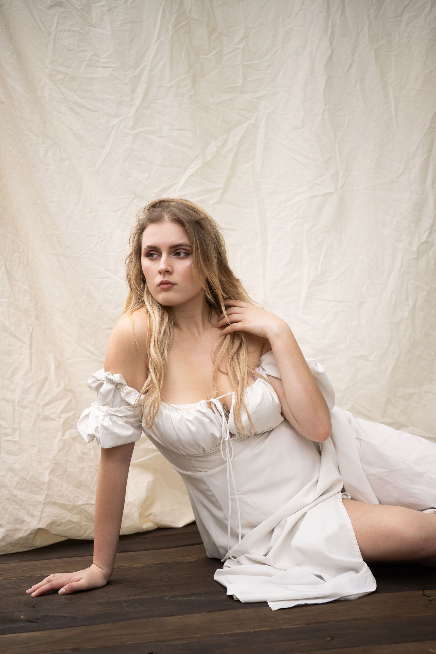 Young blonde woman in white off-shoulder dress sitting gracefully on wooden floor against cream fabric backdrop.