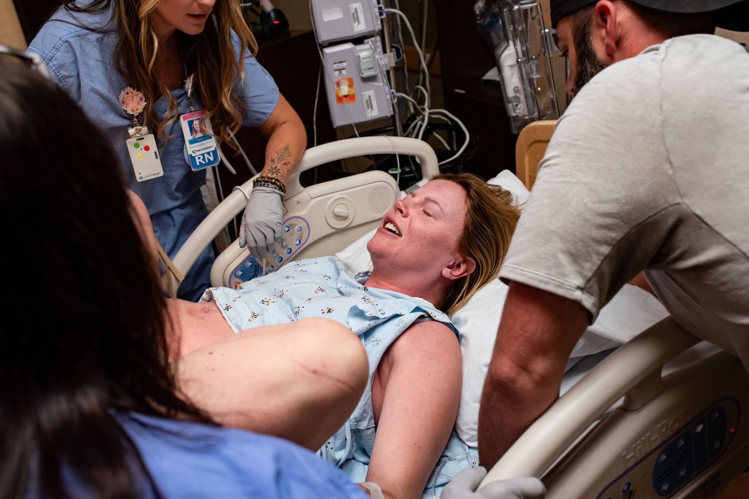 Woman in hospital bed laughing during labor, surrounded by supportive medical staff and partner in delivery room.