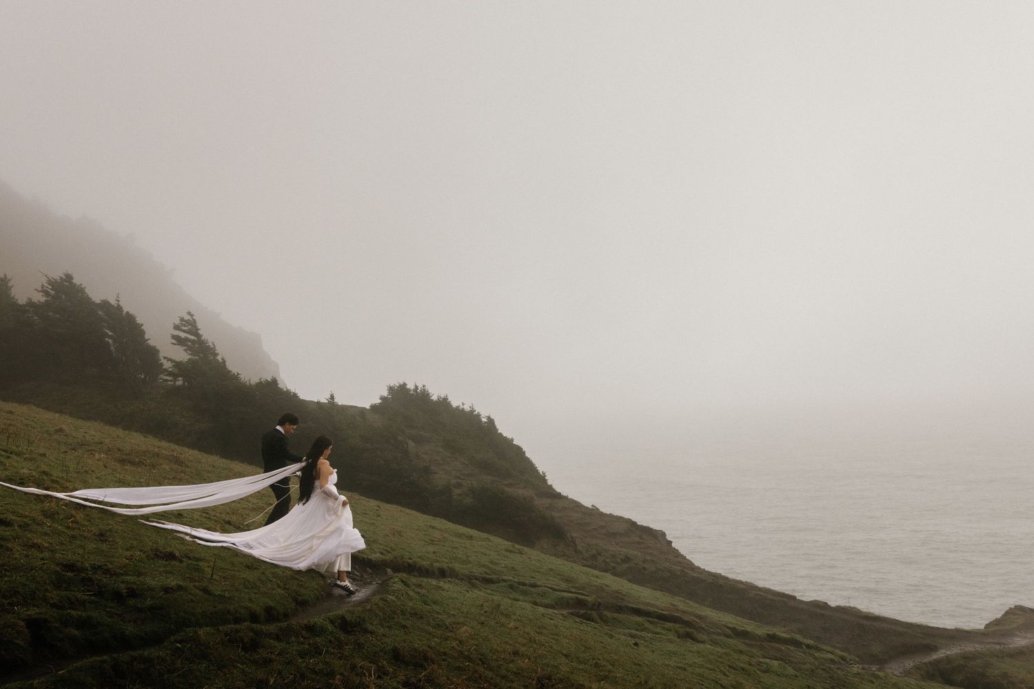 Bride in flowing white gown and veil on misty coastal hillside with groom, foggy ocean backdrop.