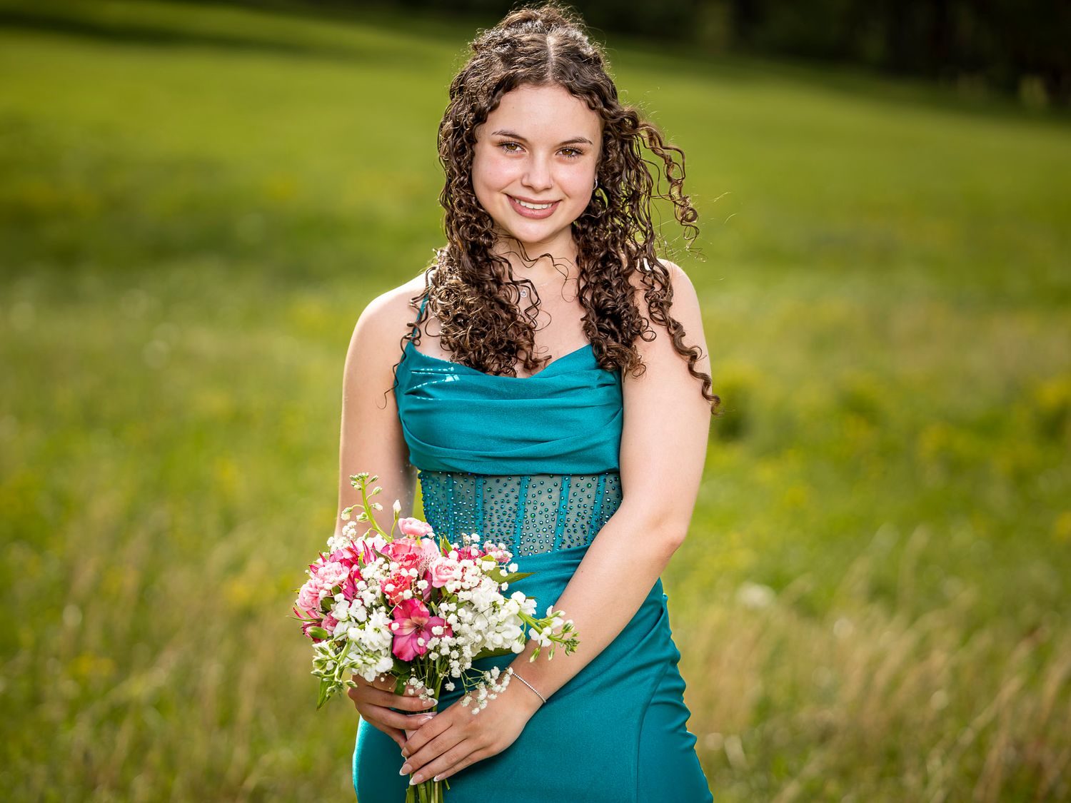 Young woman in teal dress holding pink and white flower bouquet smiling outdoors in green field.