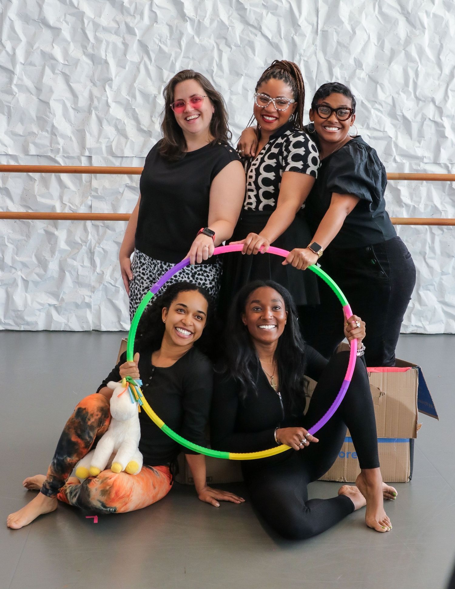 Five women smiling and posing together with a colorful hula hoop in a bright studio space.