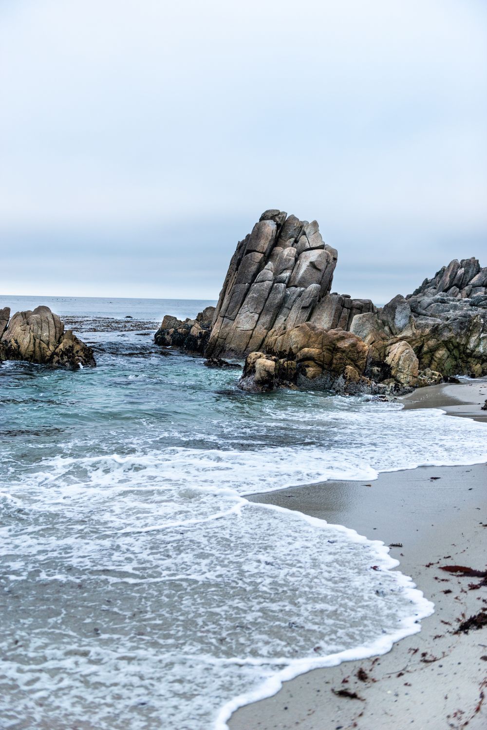 Rocky coastal shoreline with crashing waves and a dramatic stone formation rising from the Pacific Ocean on a cloudy day.