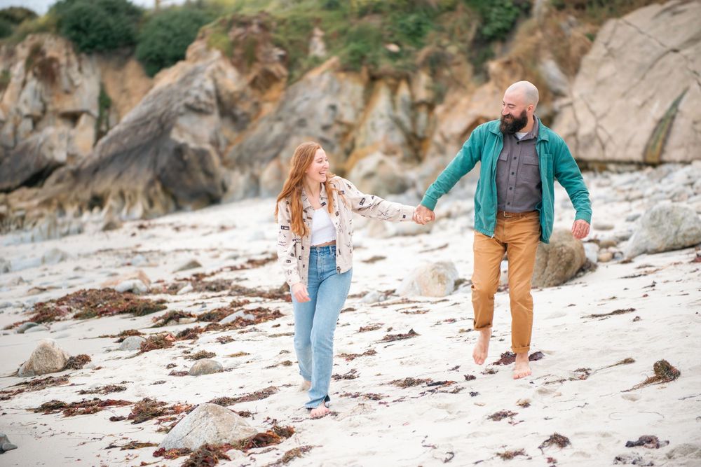 A couple holds hands and walks along a scenic rocky beach, the woman in jeans and the man in a teal jacket.
