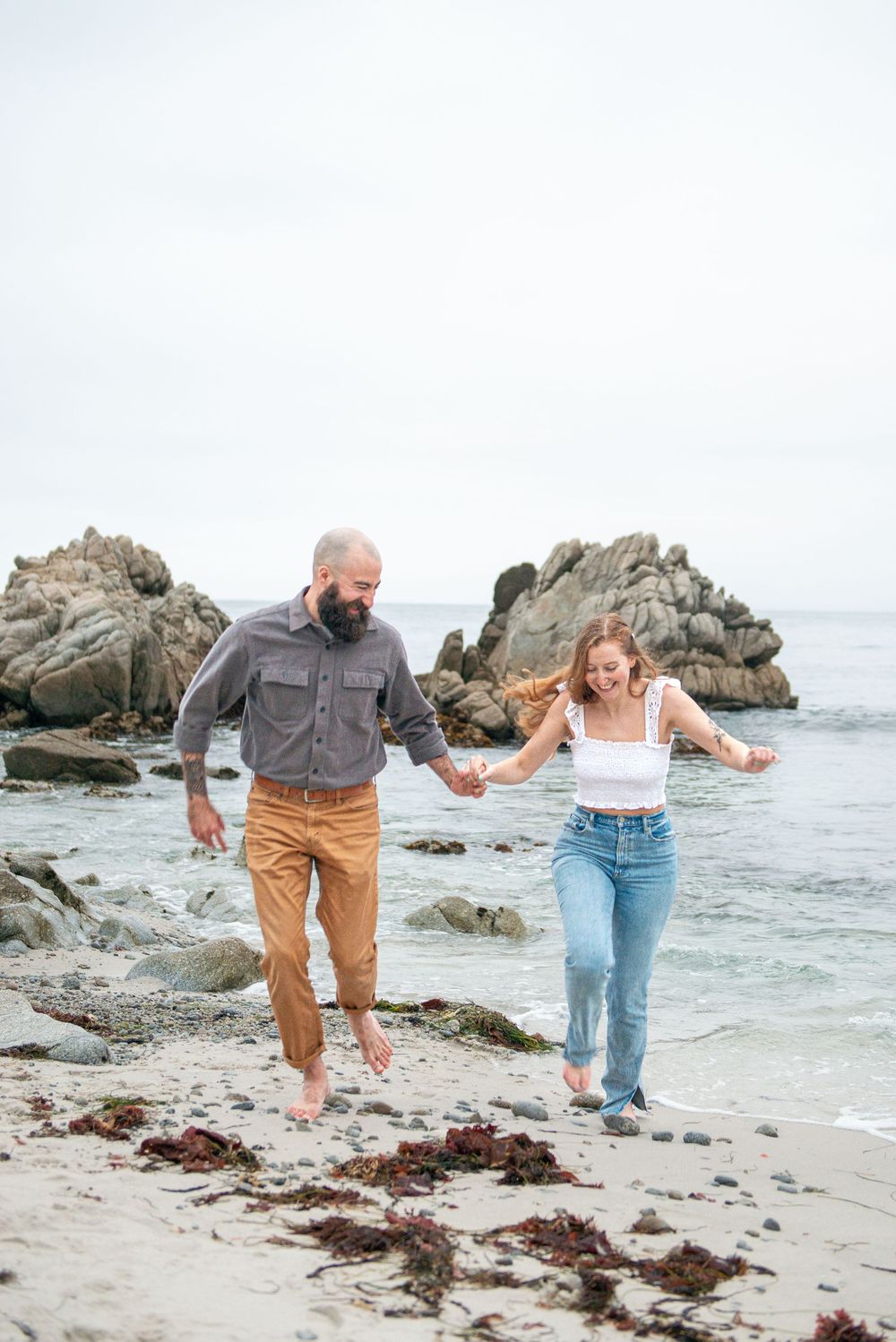 A couple holds hands and laughs while walking barefoot on a rocky beach with ocean waves behind them.