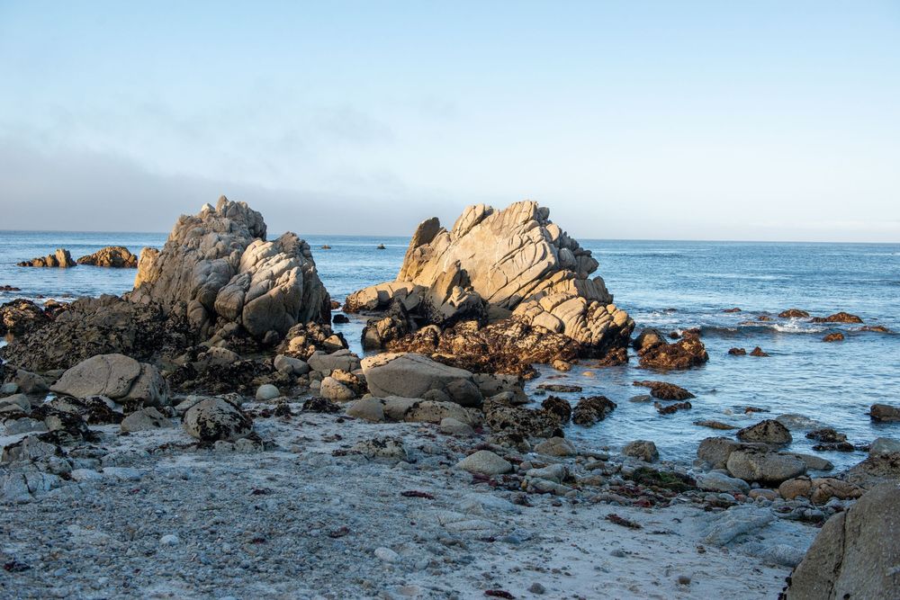 Rocky coastal shoreline with large boulders and calm blue ocean waters under a clear sky at golden hour.
