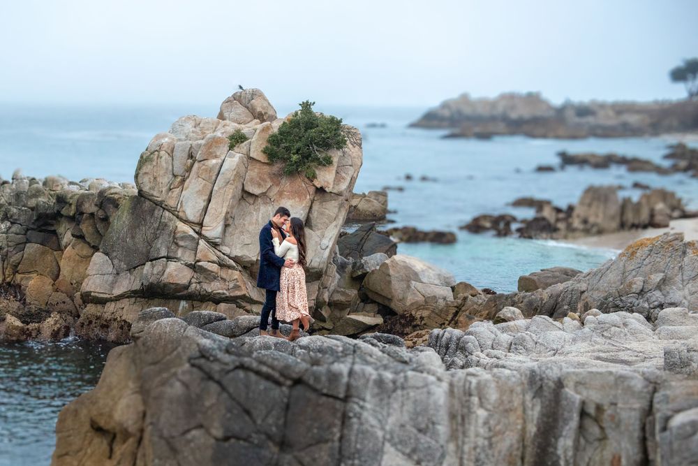 Couple embracing on rocky coastal cliffs with misty ocean waves and lush greenery in the background.