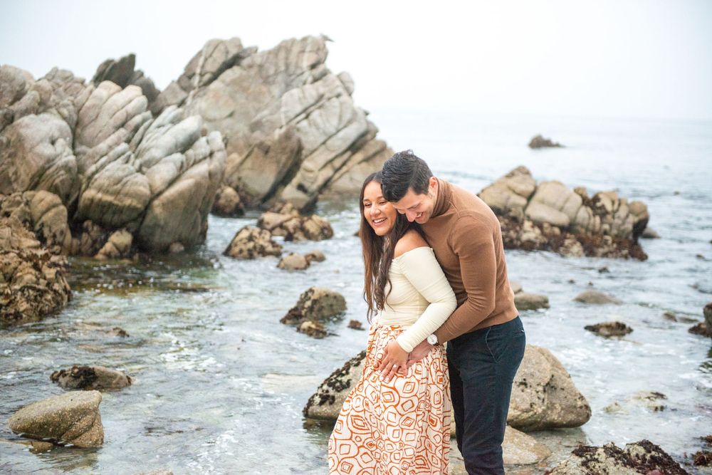 Couple embracing romantically on rocky Pacific coastline, woman in floral skirt and man in brown sweater, misty ocean backdrop.