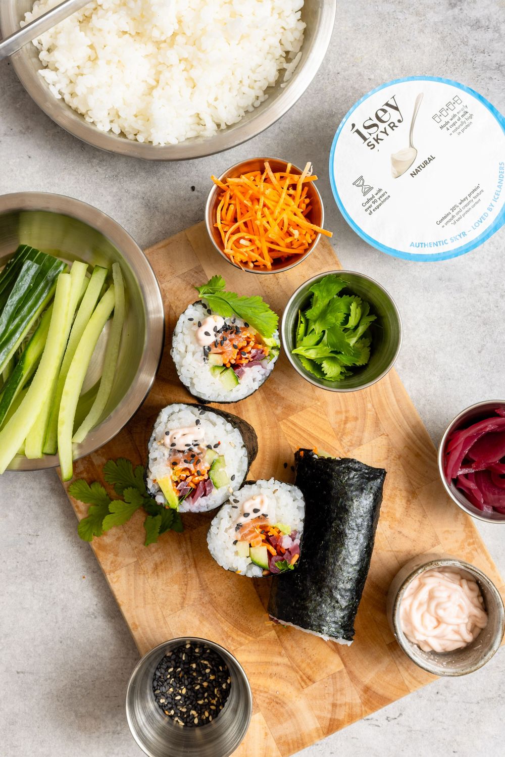 Sushi burrito prep board with rice, nori rolls, shredded carrots, cucumber, greens, and dipping sauces on a wooden cutting board.