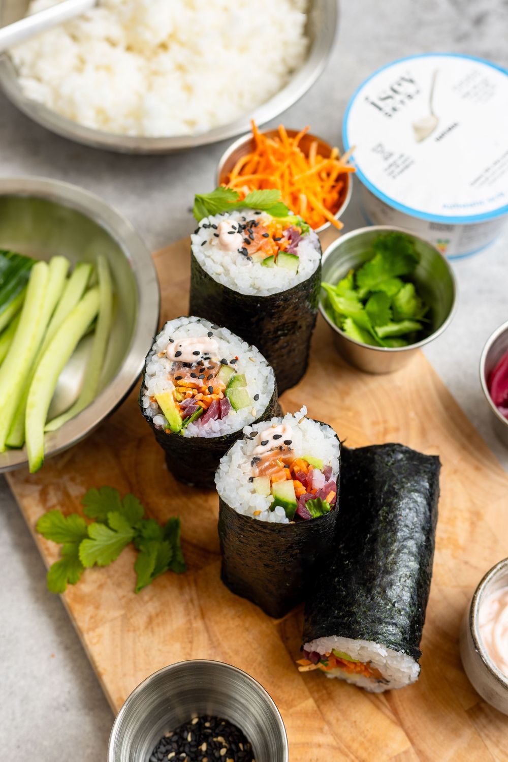 Homemade sushi rolls with rice, carrots, cucumber, and vegetables on a wooden cutting board with fresh ingredients.