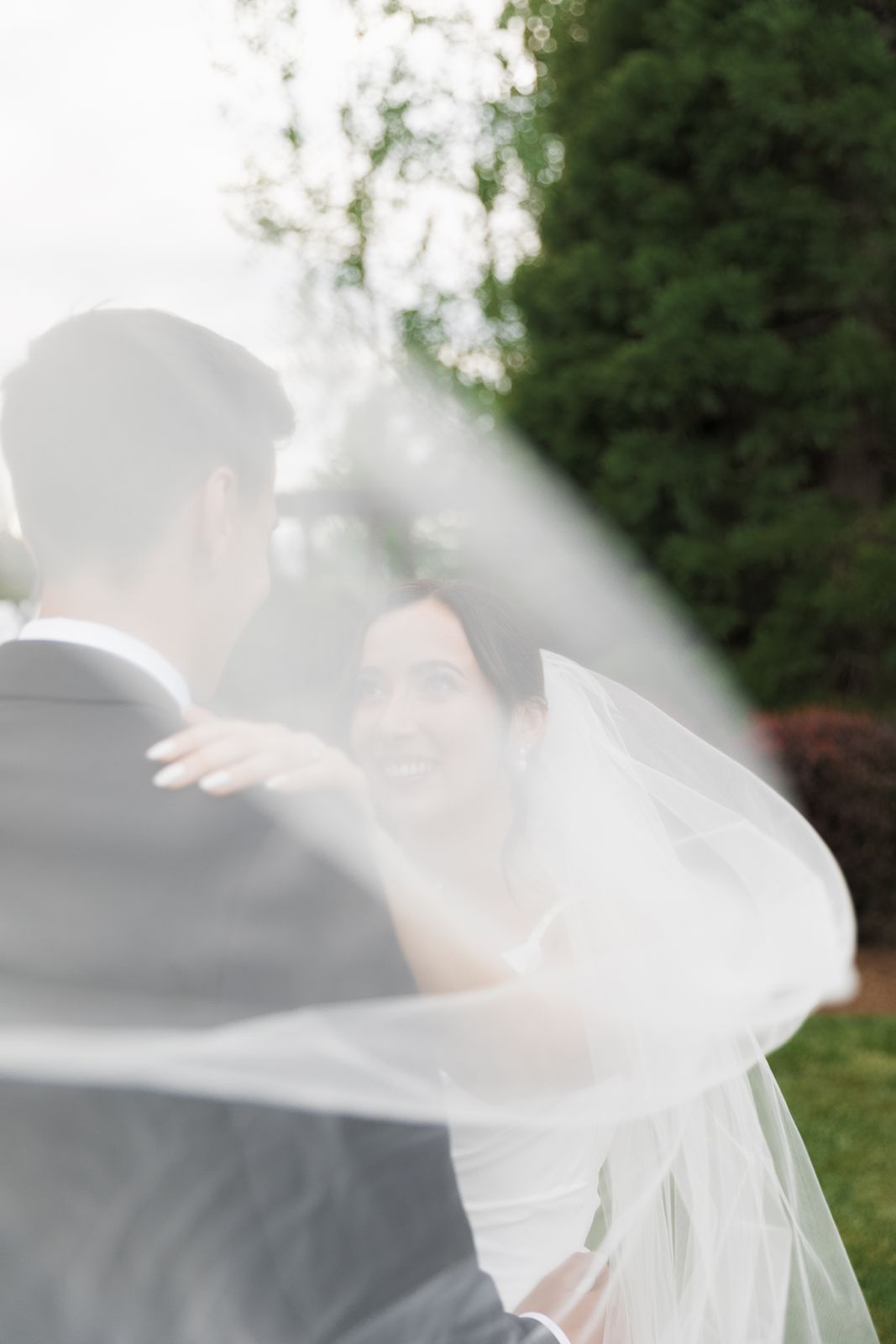 Bride smiles at groom as flowing white veil billows around them in a dreamy outdoor wedding portrait.