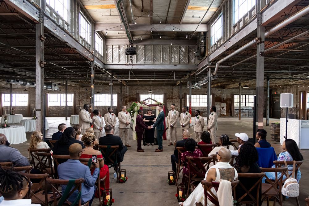 Wedding ceremony in rustic industrial warehouse venue with guests seated in wooden chairs facing altar with wedding party.
