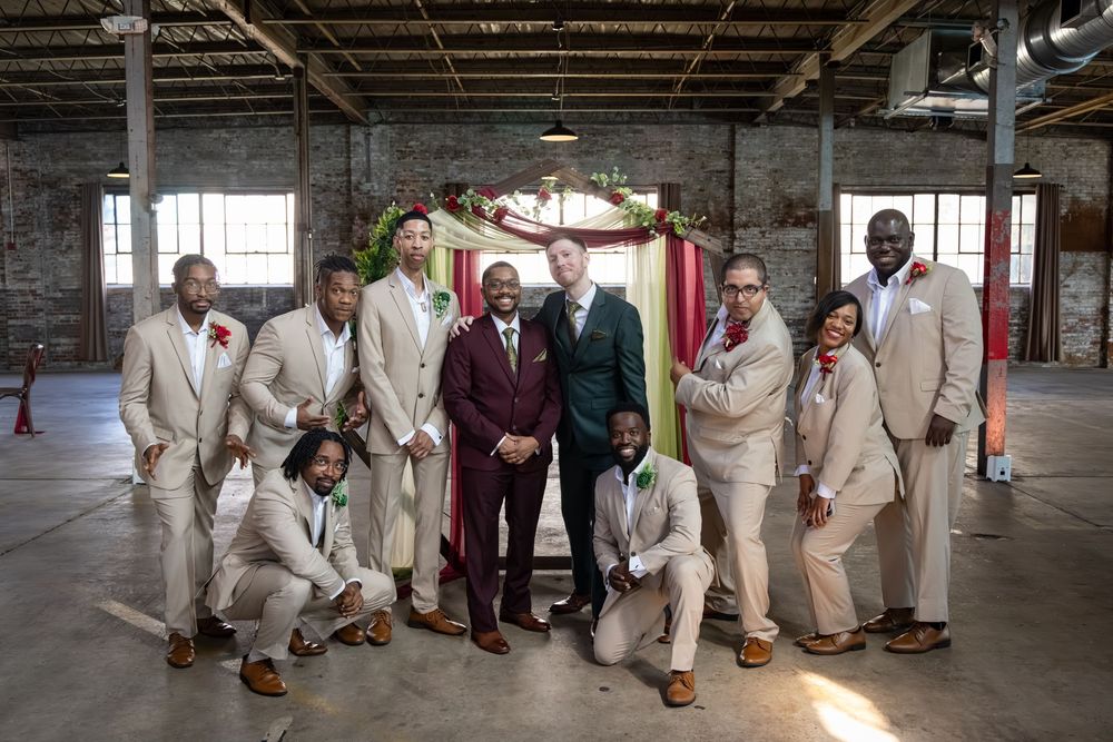 Groom and groomsmen in tan suits pose together in rustic industrial wedding venue with floral arch backdrop.