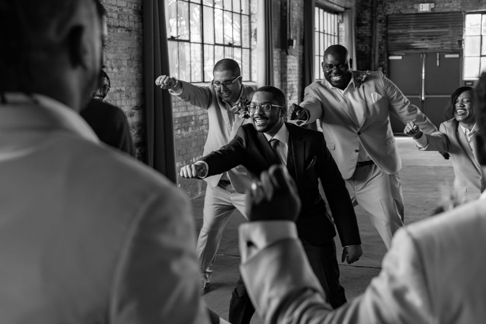 Black and white photo of groomsmen dancing joyfully at a wedding reception in a rustic brick venue.