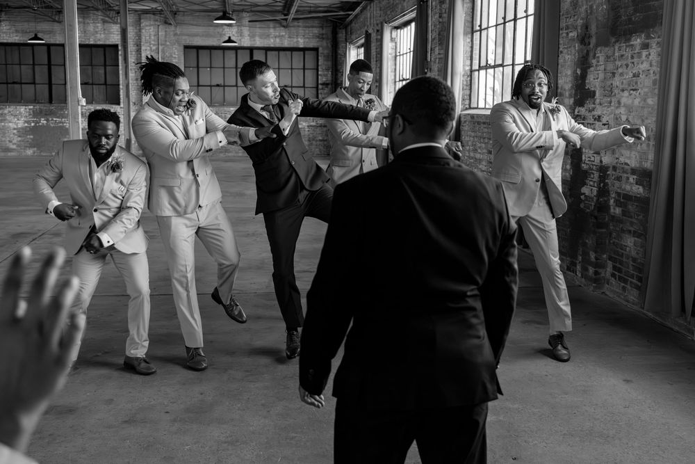 Black and white photo of groomsmen in suits dancing and celebrating together in a rustic industrial venue.
