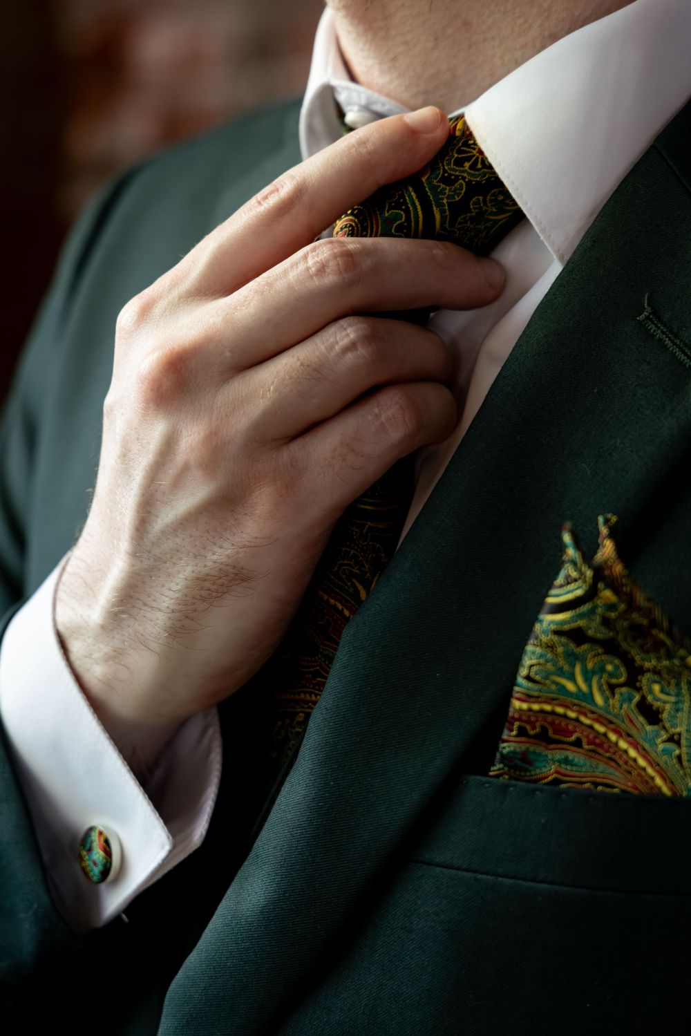 Man in green suit adjusting paisley tie with matching pocket square and gold cufflink.