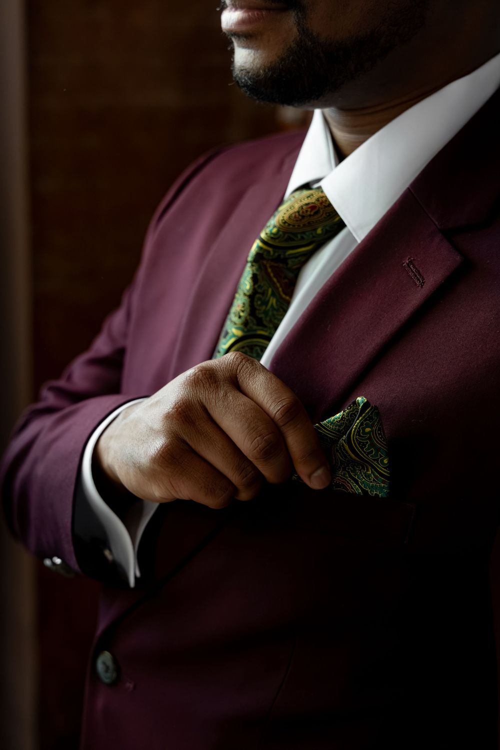 A man in a burgundy suit adjusting his green paisley tie and matching pocket square.