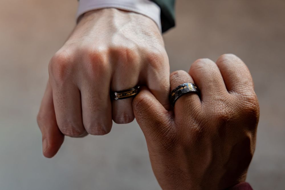 Two hands wearing matching black wedding bands touching fists, symbolizing a same-sex marriage commitment.