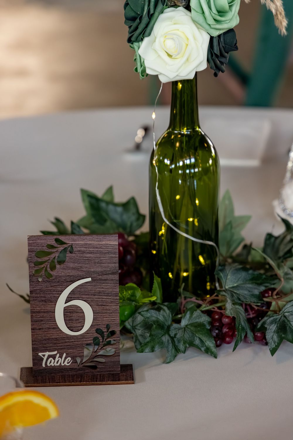 Rustic wedding Table 6 sign with wine bottle centerpiece, white roses, ivy, and fairy lights on white tablecloth.