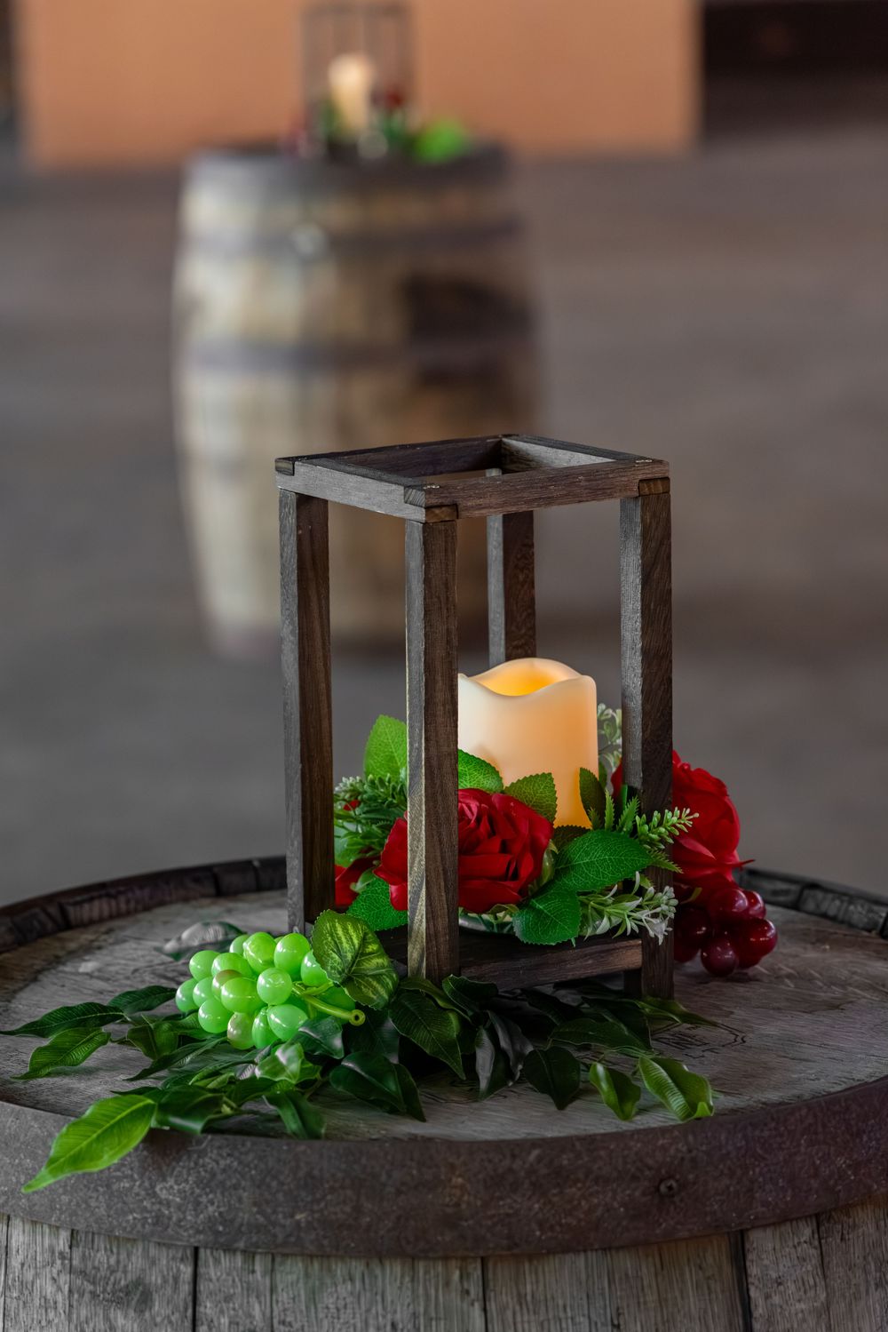 Wooden lantern with glowing candle surrounded by red roses and green grapes on a rustic wine barrel centerpiece.