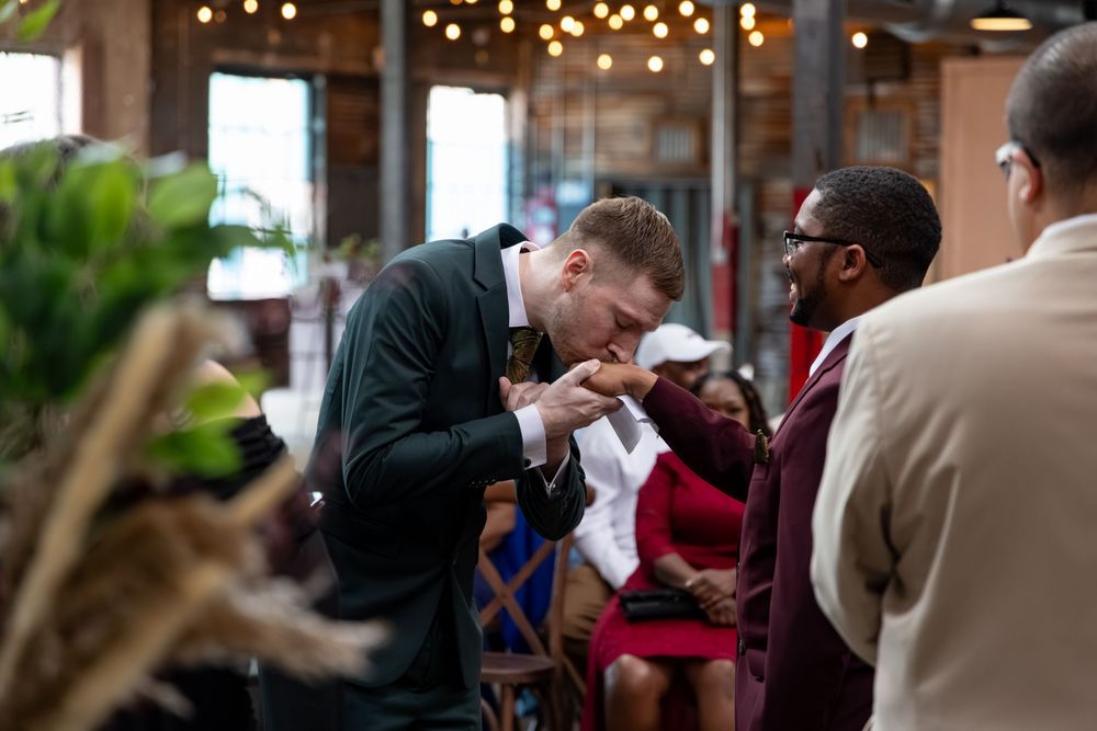 Man bowing to kiss a woman's hand at an indoor social gathering with guests in formal attire.