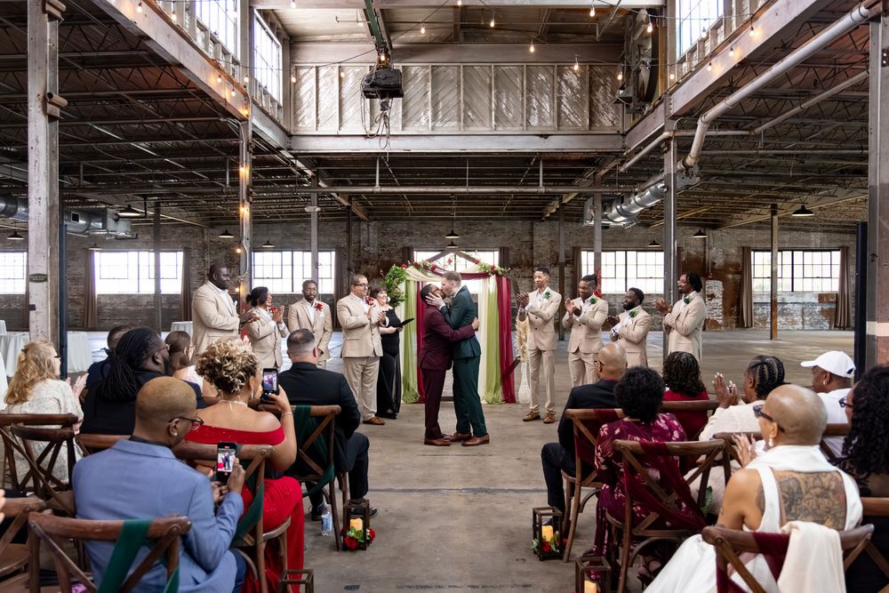 Same-sex wedding ceremony in rustic industrial venue with guests seated as couple exchanges vows before officiant.