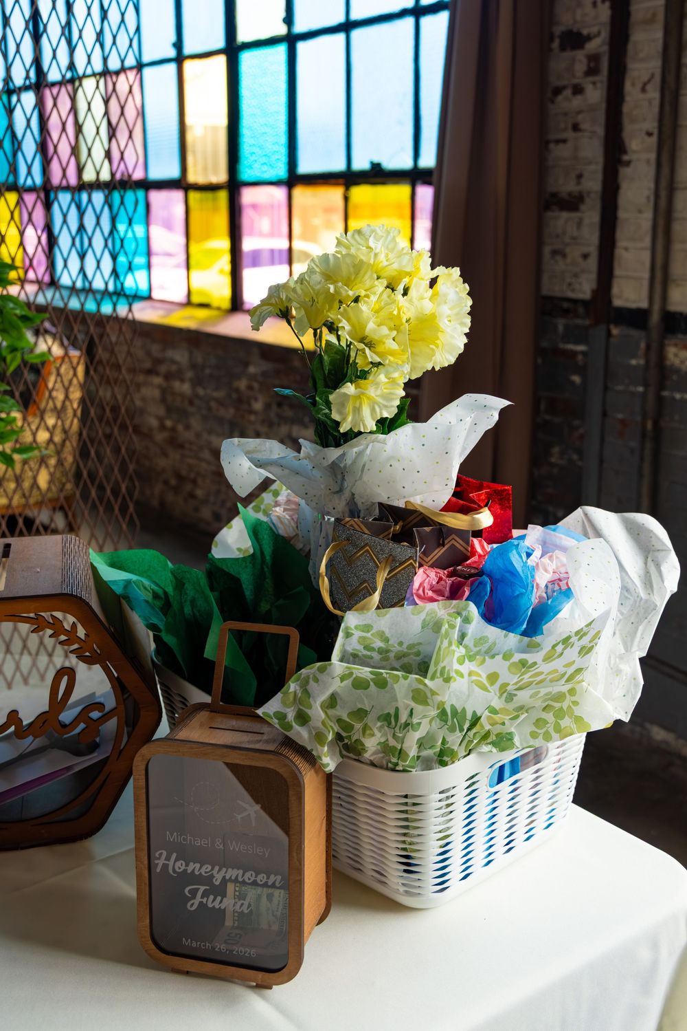 Wedding gift table with honeymoon fund sign, white basket, tissue paper, and yellow flowers near colorful stained glass window.