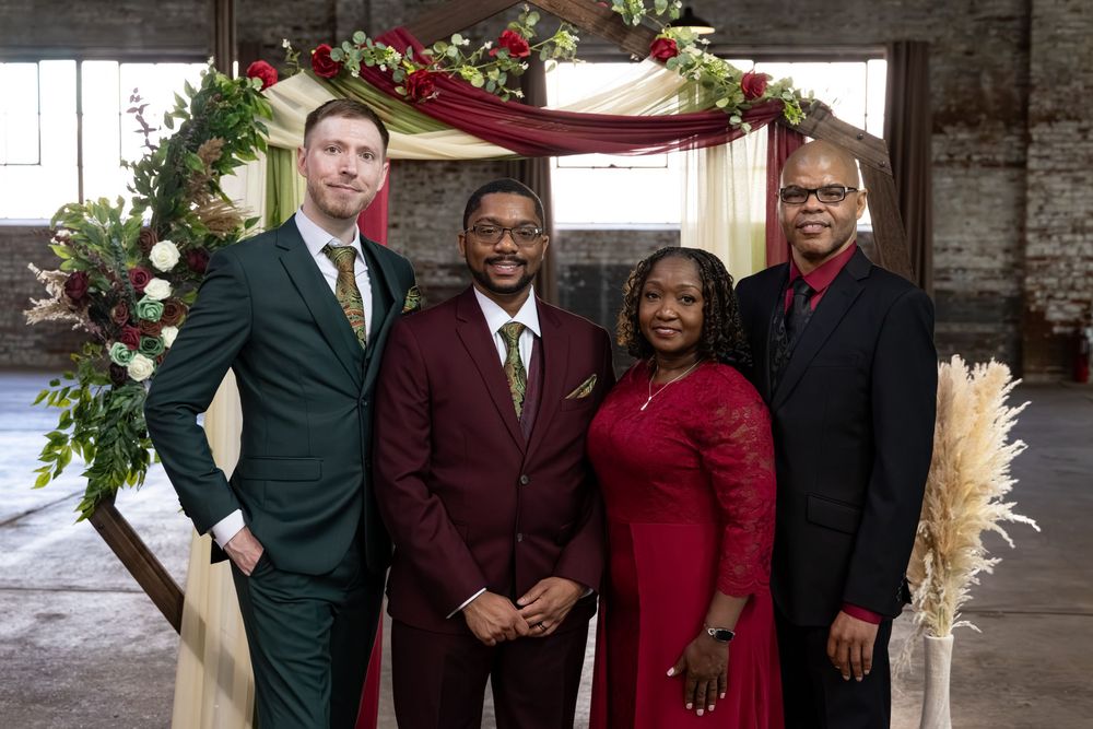 Four people pose together at wedding venue with floral arch, wearing formal attire in burgundy and green.