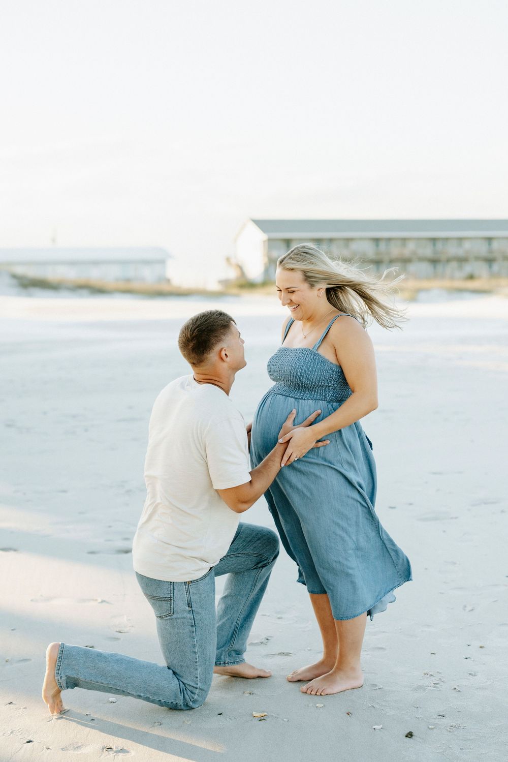 A man kneels on the beach holding a pregnant woman's belly while she smiles down at him during a maternity photoshoot.