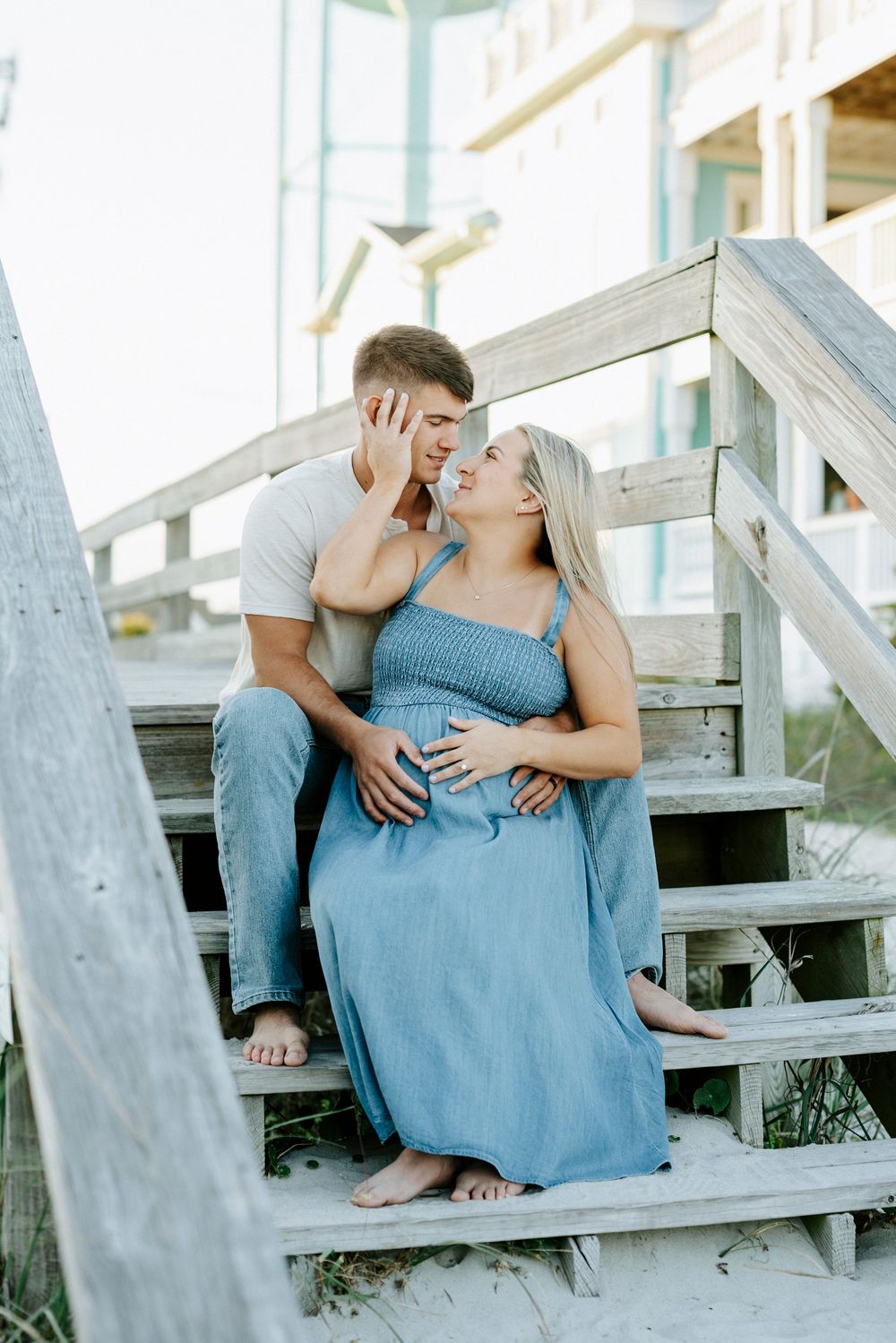 Couple sharing a tender moment on beach boardwalk stairs, pregnant woman in blue dress with partner embracing her.